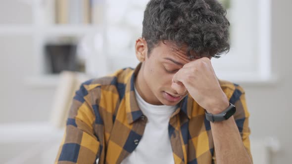 Exhausted Middle Eastern Teen Boy with Migraine Rubbing Eyes and Putting on Eyeglasses Sitting alt