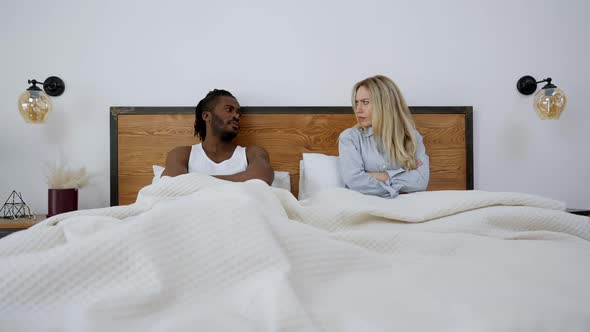 Wide Shot of Argued Interracial Couple Lying in Bed with Dissatisfied Facial Expression Looking at alt