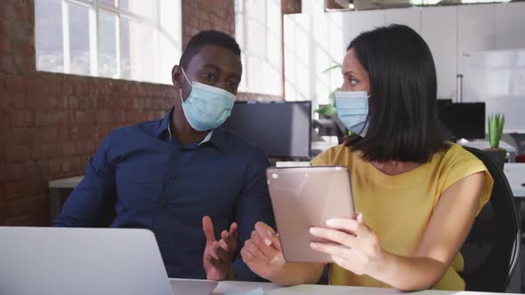 Diverse male and female business colleagues wearing face masks sitting at desk using tablet alt