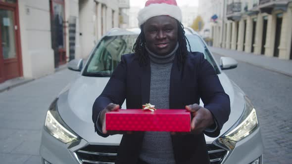 Young Black Man with Santa Hat Giving Red Wrapped Christmas Gift in Front of His Car alt