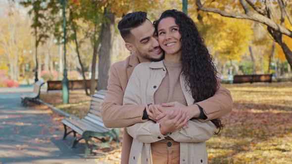 Hispanic Guy Hugs Tight Beloved Girlfriend From Behind in Autumn Park Happy Smiling Couple on Date alt