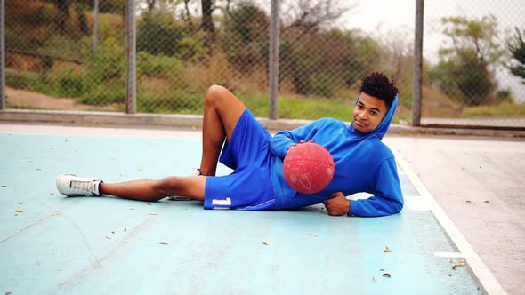 Young African American Laying on the Floor of the Playing Field and Playing with Basketball Ball alt