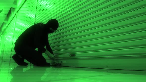 Cat burglar breaking in to a store or storage locker by picking the lock while wearing a hood and ba alt