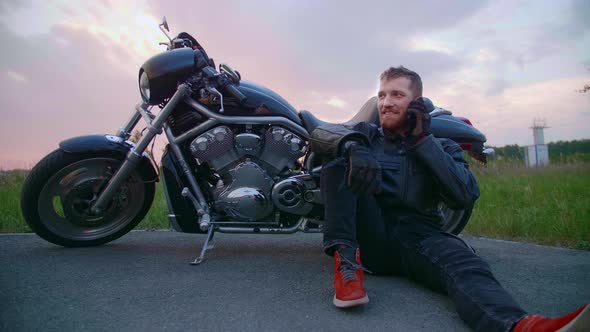 A Middleaged Male Biker Sits By a Motorcycle and Talks on His Mobile Phone