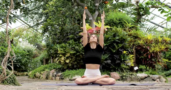 A Young Brunette Woman is Doing Yoga Exercises in the Lotus Position on a Mat in a Botanical Garden