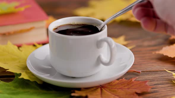 Hand Stirring Coffe with Spoon in Cup on Wooden Table with Book and Colorful Autumnal Maple Leaves