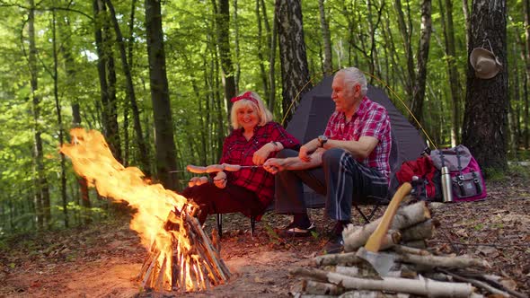 Senior Elderly Grandmother Grandfather Cooking Frying Sausages Over Campfire in Wood at Camping alt