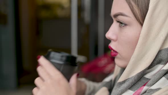 woman straightens her scarf on her head and drinks coffee from a disposable cup alt