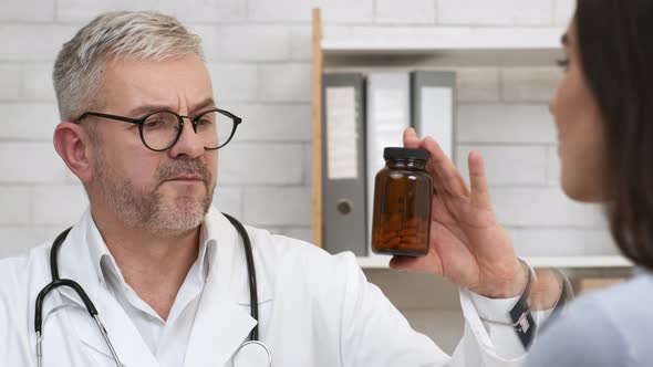 Male Doctor Prescribing Medication Showing Medicine Bottle To Patient Indoors alt