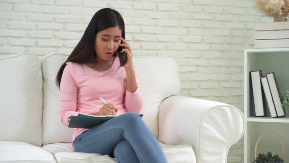 Young Asian woman writing notebook and talking smartphone on the couch in the cozy living room.