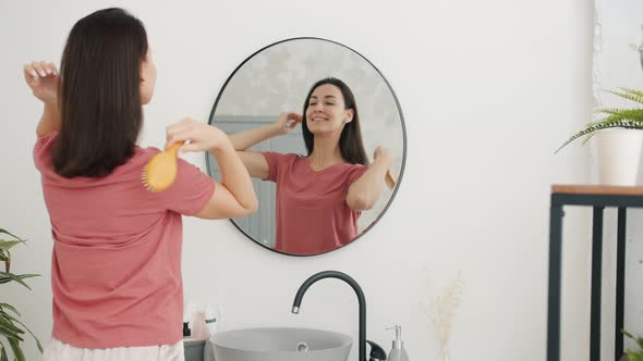 Portrait of Cute Young Woman Busy with Beauty Routine Brushing Hair in Bathroom alt