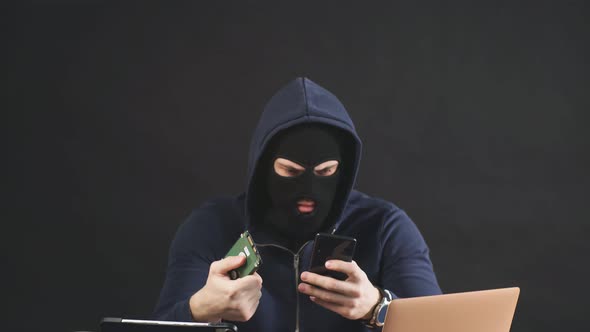 Male Hacker in Black Mask and Hood Uses Laptop To Hack a Hard Drive. alt
