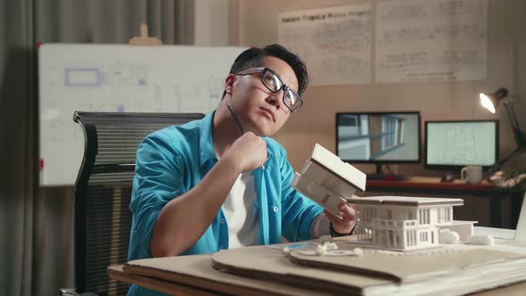 Asian Man Engineer With A Laptop Thinking While Holding Paper Model Of House At The Office alt