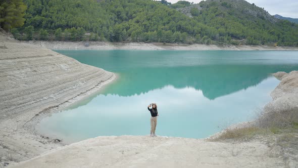 Girl Stands By Turquoise Lake and Takes Selfie with Phone Wide Wiew alt