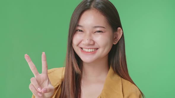 Smiling Young Asian Woman Showing Gesture Peace While Standing On Green Screen In The Studio alt