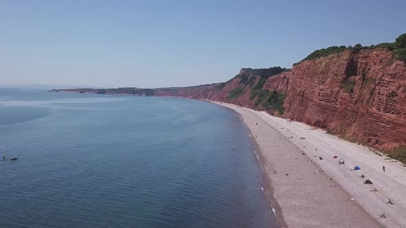 Wide shot of the red cliffs at Budleigh Salterton, Devon. Tide is in, and holiday makers are on the alt