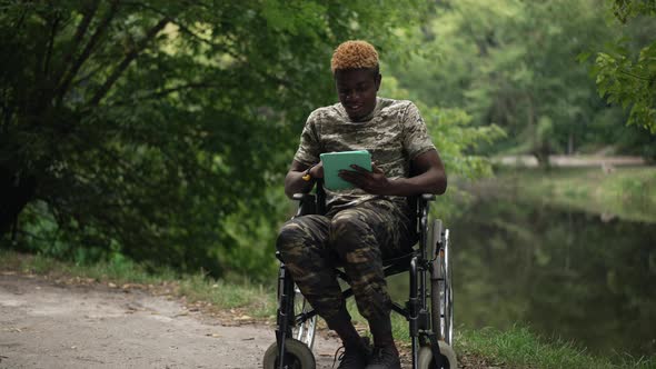 Smiling African American Man in Wheelchair Surfing Social Media on Tablet Sitting on Lake Shore in alt