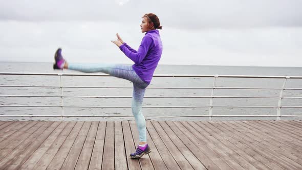 Young Woman Exercising Kicking Legs and Stretching on the Beach By the Ocean Early in the Morning alt