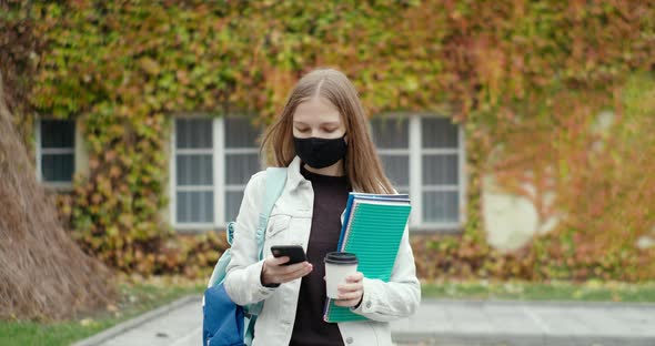 Female Student in Mask Uses Smartphone Mobile App and Walks Outside with Coffee
