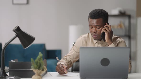 Adult Black Man is Communicating By Mobile Phone Sitting at Table with Laptop Writing Notes Working alt