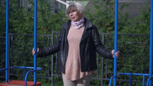 Portrait of Grieving Sad Senior Woman Standing on Playground at Swings Thinking alt