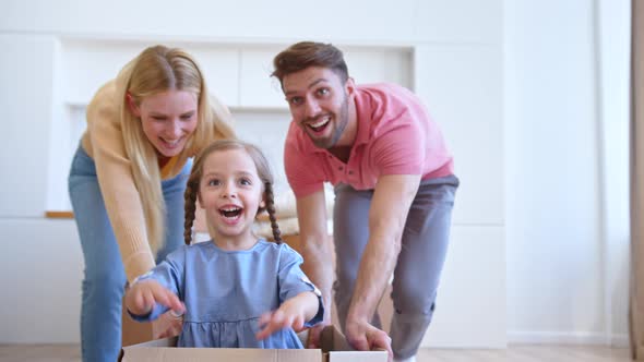 Joyful mother and father move brown cardboard box with funny little preschooler daughter alt