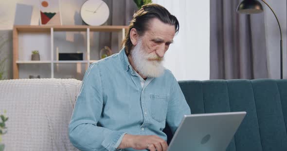 Senior Bearded Man Working on Laptop at Home and Looking at Camera with Thumb Up alt