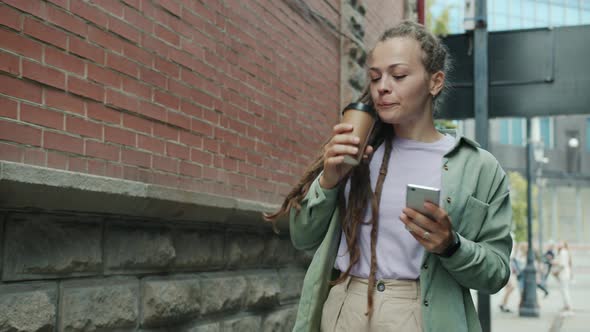 Beautiful Girl with Dreads Drinking to Go Coffee and Using Smart Phone Walking Outside Alone alt