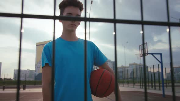 Cinematic Shot Portrait of Serious Teenager with Basketball Ball on Basketball Court at Sunset Sad