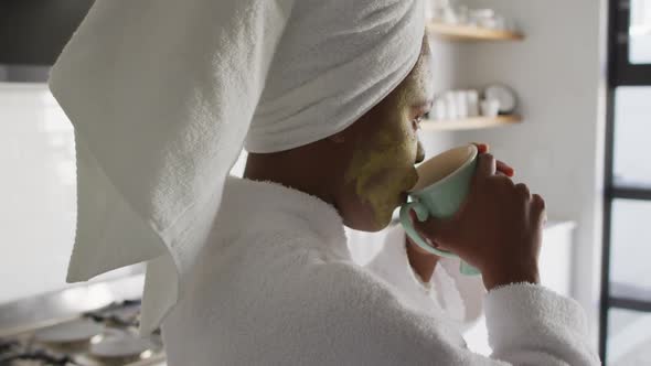 Happy african american woman with beauty mask on face, drinking coffee in kitchen alt
