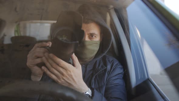 A Man in a Black Hoodie and a Protective Medical Mask on His Face Sitting Behind the Wheel of a Car alt