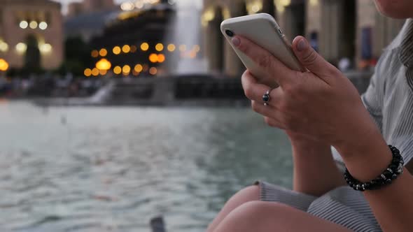 A Woman Sitting in a Pond in a Public City Park Uses a Smartphone Holding in Her Hands and Touches