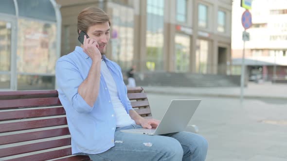 Man Talking on Phone and Using Laptop While Sitting Outdoor on Bench alt