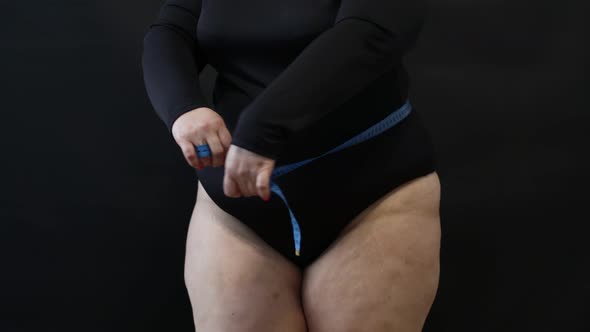 a Fat Woman with Cellulite Measures Herself with a Measuring Tape on a Black Background