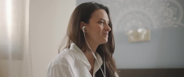 Close up of a young woman talking to someone through earphones while sitting on bed.  alt