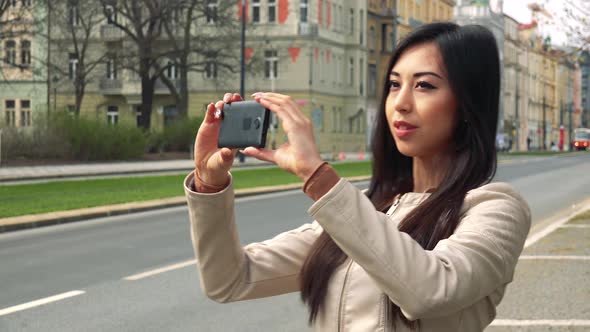 A Young Asian Woman Takes Pictures of Her Surroundings with a Smartphone in a Street in Urban Area alt