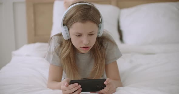 Joyful Teen Girl Is Reading Text on Screen of Mobile Phone, Lying on Bed, Using Wireless Headphones alt