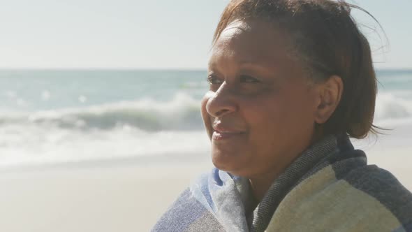 Smiling senior african american woman with blanket on sunny beach alt