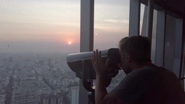 Traveler Man Watching Through the Stationary Binoculars at a Scenic Overlook at Sunset in Ho Chi