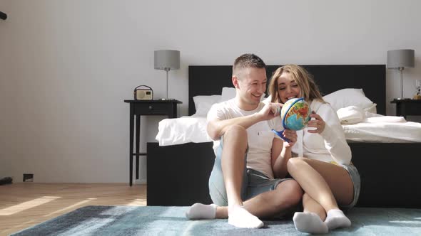 Young Couple Sitting Beside the Bed Are Looking at Globe Choosing a Place To Travel on Honeymoon alt