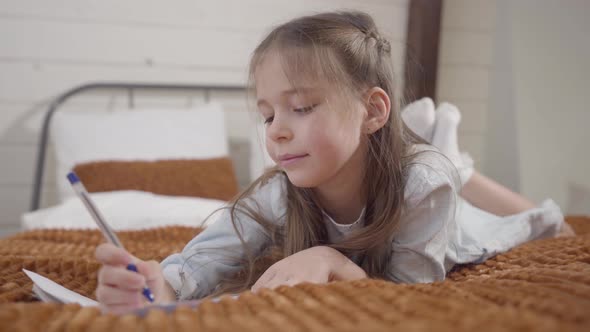 Portrait of Charming Caucasian Girl Lying on Bed and Writing. Intelligent Child Showing i Got an alt