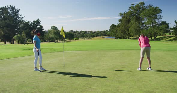 Two caucasian women playing golf wearing face masks one taking shot from bunker alt