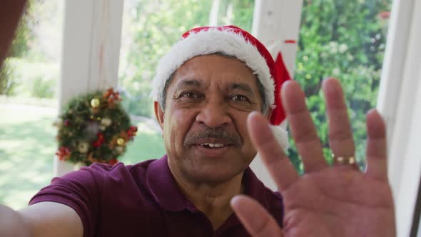 Portrait of happy senior man in santa hat waving hand during christmas video call alt