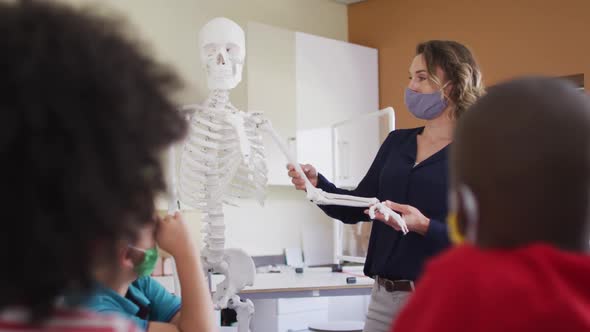Female teacher wearing face mask using human skeleton model to teach students in class alt