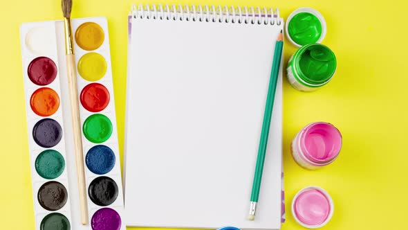 a Variety of Watercolor Paints in Jars with a White Sketchbook on a Pastel Yellow Background alt