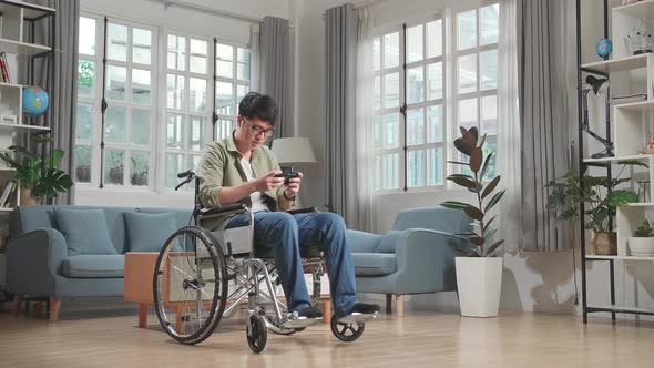 Asian Man Sitting In A Wheelchair While Use Mobile Phone Playing Video Game At Home alt