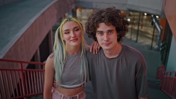 Young Millennial Couple with Unusual Appearance Smiling to Camera Embracing Against Underground alt