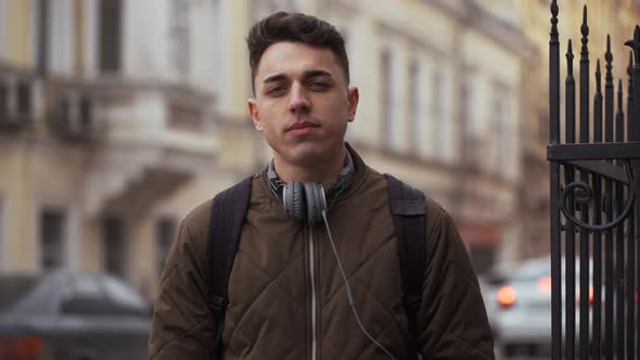 Portrait of European Guy 20s Wearing Jacket and Backpack Inhaling Fresh Air During Walk Through City alt