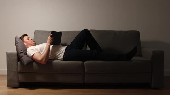 Young adult man lies on a sofa and reads a book in the evening