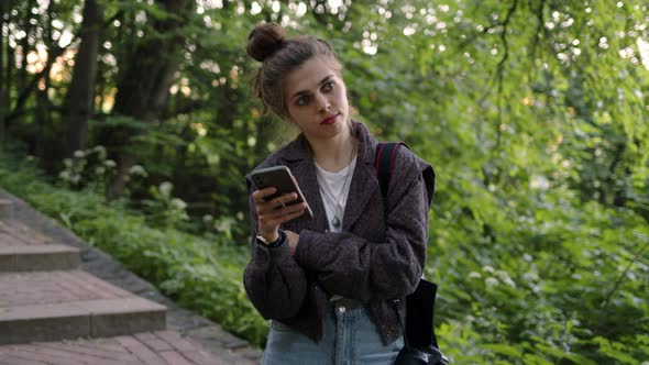 Portrait of Woman in Gray Jacket Stands with Phone in Her Hand in Park and Thinks Looks Away Then alt